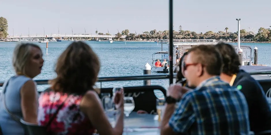 People enjoying drinks at a waterfront venue, overlooking calm blue water, boats and a bridge, with a relaxed outdoor dining atmosphere on a clear day.